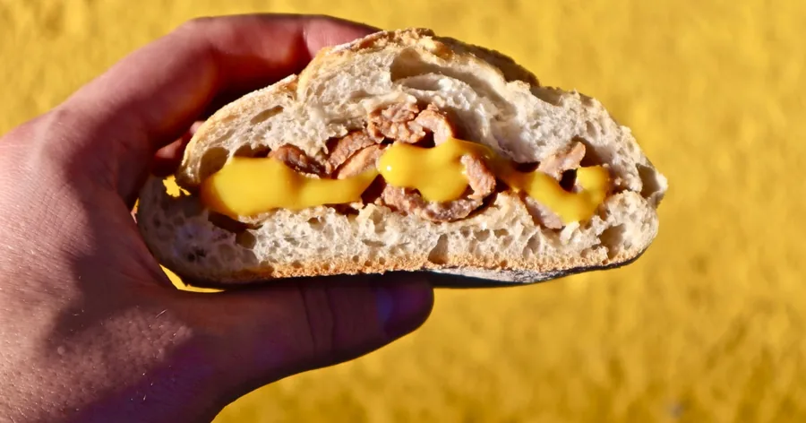 Eine Hand hält eine Bifana, ein knuspriges Brötchen mit mariniertem Schweinefleisch, vor einer gelben Wand
