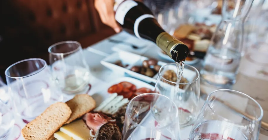 Aus einer Flasche wird Weißwein in Gläser gegossen, daneben ein Brett mit Käse und Wurst